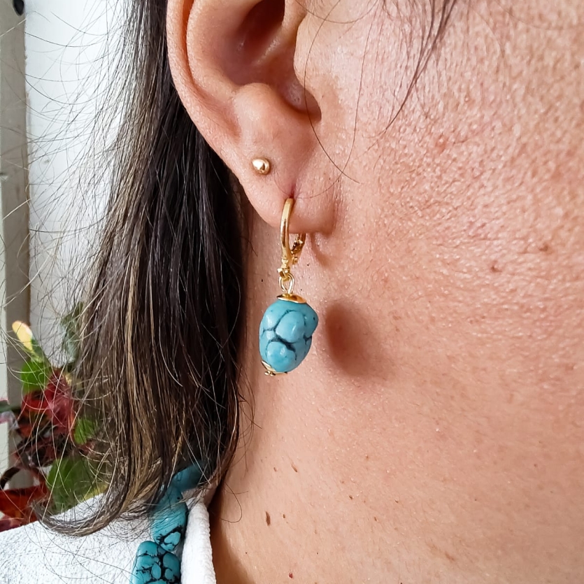 Brinco dourado pequeno com pedra turquesa pendente, usado na orelha, acompanhado por colar de pedras turquesa.