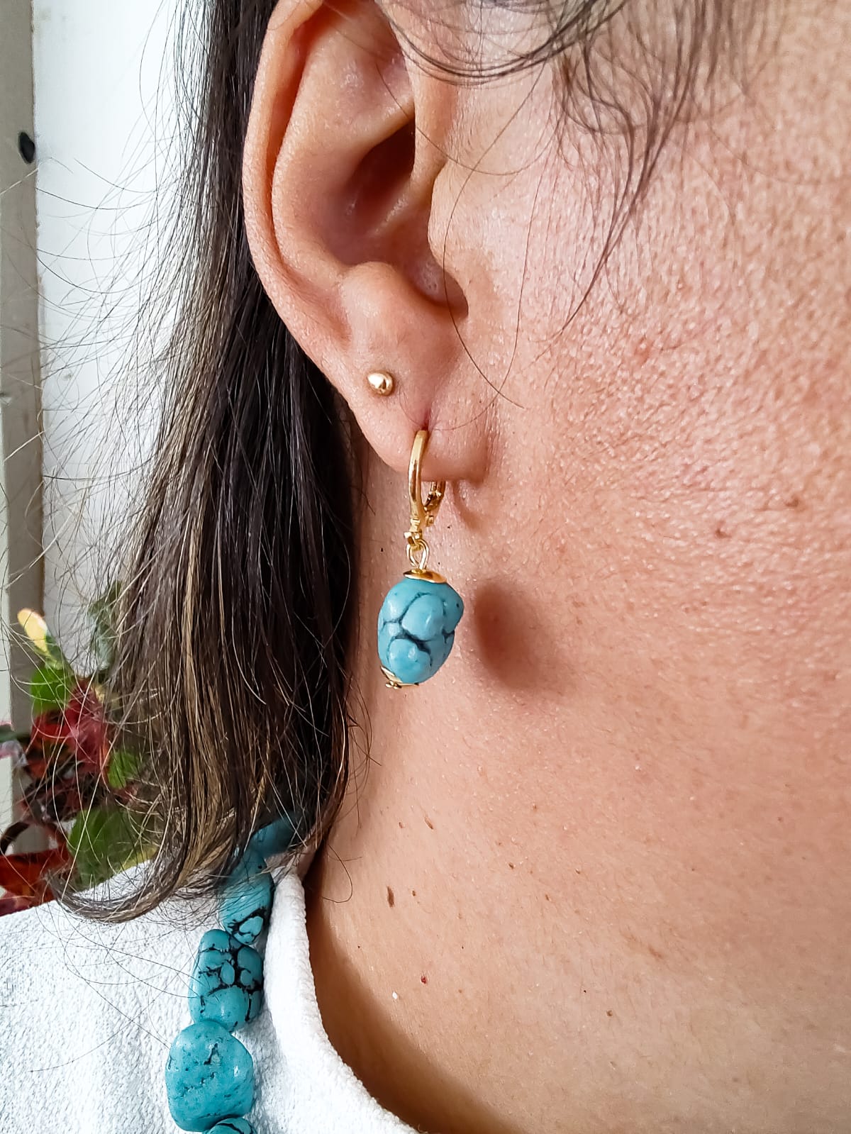 Brinco dourado pequeno com pedra turquesa pendente, usado na orelha, acompanhado por colar de pedras turquesa.